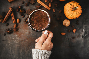 Top view of female hand holding mug of hot chocolate, pumpkins and spices on rustic background, cozy autumn scene