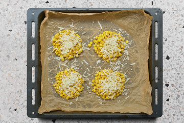 Top-down view of a baking sheet with parchment paper. Corn rounds are topped with grated cheese and arranged neatly. The sheet sits on a stone countertop.