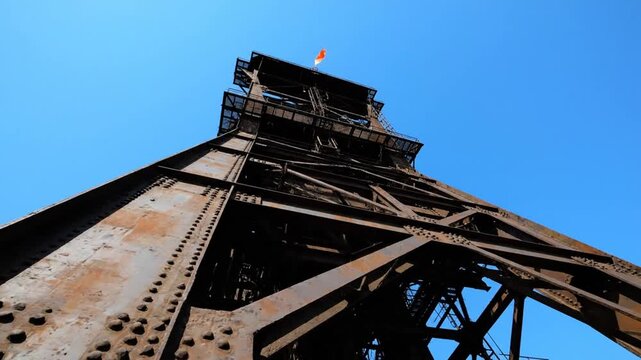 Low-angle view of a tall, rusty industrial mining tower against a clear blue sky, with a small flag at its peak.