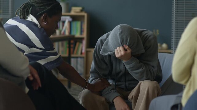 Depressed gen Z Caucasian guy surrounded by multiethnic peers sitting in circle in classroom supporting him and holding hands during group therapy session with counselor