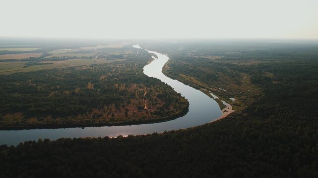 Aerial view of river bend flowing through valley among meadows and forests with slight haze on horizon. Concept of silence and pacification
