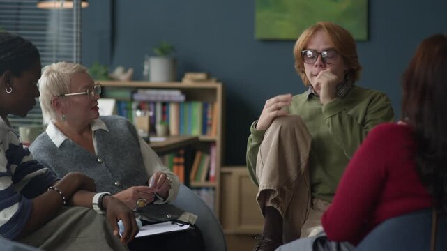 Medium long shot of young Caucasian red-haired student talking in front of multiethnic peers listening to him attentively during group therapy session in modern office with mature female counselor