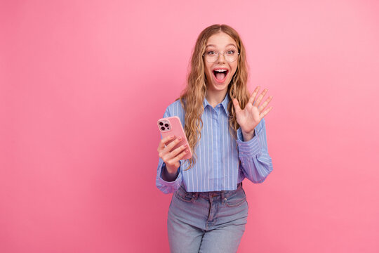 Excited young woman waving while holding a smartphone against a pink background, sharing enthusiasm with a lively and cheerful expression