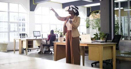 Donning VR headset, African American businesswoman exploring VR with controller at desk, copy space - Powered by Adobe