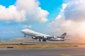 A large cargo plane takes off from the runway at an airport. The sky is blue with some scattered clouds. The sun shines brightly on the side of the aircraft and surrounding terrain