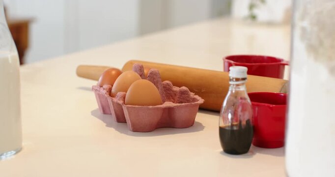 Camera panning left, showing egg carton, rolling pin, vanilla, flour jar and milk bottle on counter