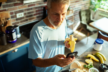 Man eating banana and using smartphone in kitchen