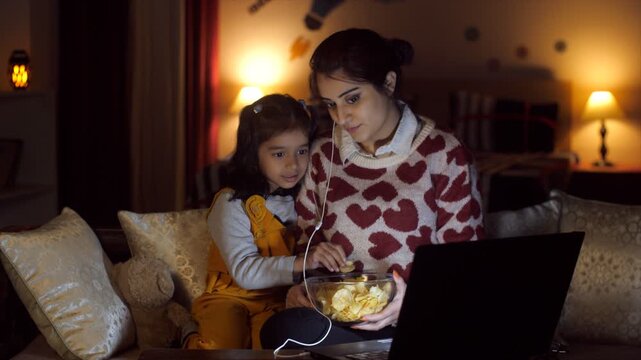 A cheerful mother-daughter watching an online movie - OTT  leisure time  late-night weekend movie plan  parenting  single mother. Pretty female and her cute kid eating potato chips while watching a...