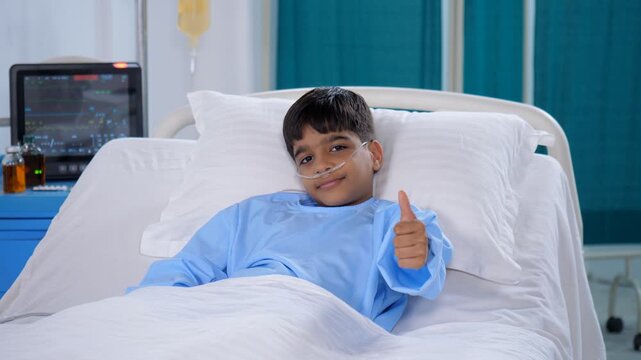 A young boy patient is posing for the camera in the hospital with a thumbs-up gesture - wellness  cheerful. A small boy wearing blue hospital clothes is admitted to the hospital - sick patient  med...