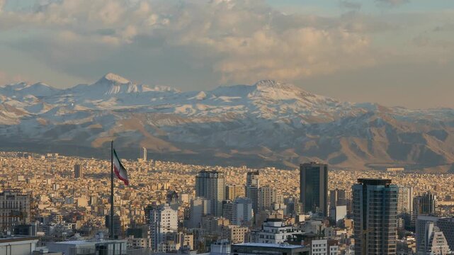 Daylight view of Tehran city with the Iranian flag in the foreground and the snow-covered Alborz mountains in the background, blending urban skyline with majestic nature.