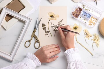 Woman putting dry flowers onto paper sheet at white marble table, top view