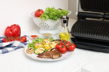Electric grill, tasty meat, vegetables, mushrooms and spices on white table in kitchen, closeup