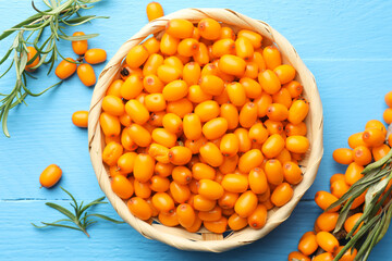 Fresh sea buckthorn berries in wicker bowl and leaves on light blue wooden table, flat lay