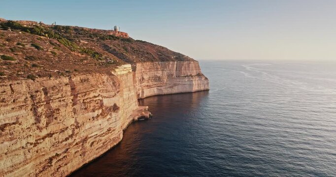 Malta: Golden sunset light reflecting on Dingli rocks cliffs, the highest point of Malta, with a radar dome on top and the Mediterranean Sea calm waters shining below, aerial drone flight footage