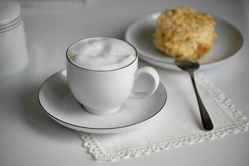 A cup of hot cappuccino with thick milk foam, served on a white table with a lace napkin. Next to it on a plate is a piece of fresh pastry.