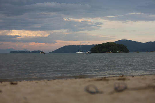 Praia tranquila com barcos ao fundo e ilhas no horizonte sob c&eacute;u nublado no fim do dia.