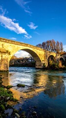 Obraz premium Stone arch bridge spans river, reflecting golden sunlight on calm water under a vibrant blue sky with wispy clouds
