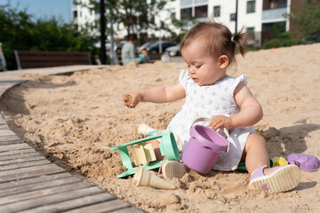 Naklejka premium adorable toddler playing with sand toys on a sunny day in a urban playground surrounded by lush greenery and modern apartment buildings, bliss, interaction, development, relax, peaceful, beige