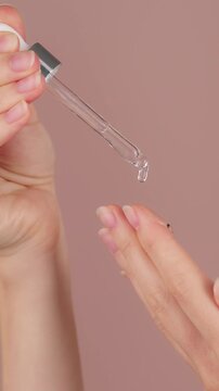 Vertical close-up of female hands on isolated pink background holding dropper, anti-aging facial serum, booster dripping on palm. Skincare, moisturizing, vitamin c, face lifting, anti-acne advertising