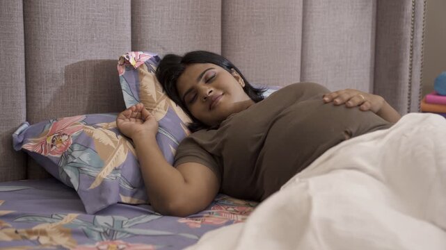 An Asian pregnant lady rubbing her belly while sleeping peacefully on the bed - sleeping during pregnancy . A young expecting Indian mother with pregnant belly - relaxation  improved sleep  relieve...
