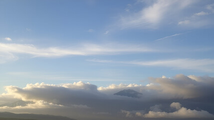 Obraz premium Serene cloudy sky with soft, feathery clouds gently embracing the majestic mountains, bathing the scene in a warm, comforting light, exuding a sense of tranquility and relaxation.