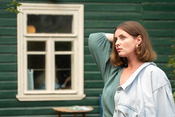 Fototapeta premium A woman in a green dress with a light denim jacket poses near a rustic green wooden house in Peredelkino