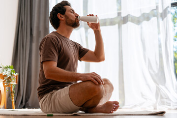 Relaxed bearded man sitting cross legged drinking from white bottle indoors in living room