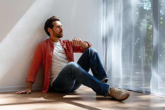 Thoughtful serious Latin man sitting on floor by window light holding eyeglasses looking away