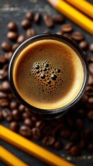 Topdown shot of a glass of espresso surrounded by coffee beans and yellow pencils on a dark surface, creating a warm and inviting atmosphere for work or study