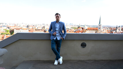 Man in blue blazer and jeans leaning on terrace railing with panoramic view of historic city and red rooftops, symbolizing lifestyle, travel inspiration, and urban culture.