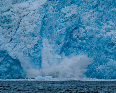 Aialik Glacier face calving ice bergs into the bay with seabirds flying in front of the mountainous spray of the splash provides perspective on its size