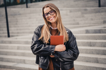 Fototapeta premium Young woman in a leather jacket holding a book, smiling confidently outdoors on city steps during a pleasant day
