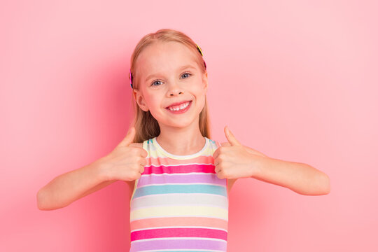 Fototapeta Adorable young girl showing thumbs up with cheerful expression on pink background wearing colorful striped dress