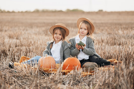 Two girls sit together in a pumpkin field wearing hats and cozy sweaters during autumn