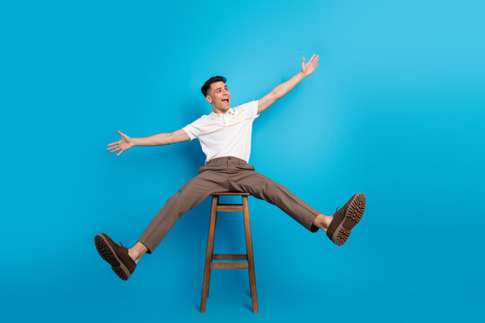 Energetic young man posing stylishly on stool with spread arms against bright blue background