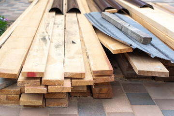 Stacked wooden planks and building materials, including metal roofing elements, laying on a patio with paving stones, ready for a home construction or repair project