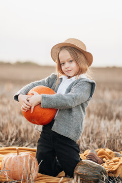 Young girl holds a large pumpkin in a field during autumn at sunset, dressed in cozy attire with a straw hat