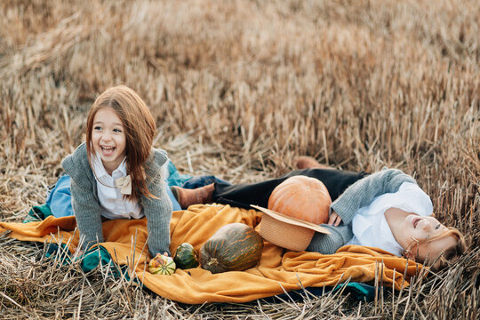 Two children laughing on a blanket in a field during autumn with pumpkins and gourds around them