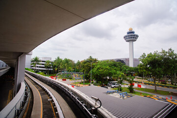 A view of an airport control tower with a monorail track in the foreground, surrounded by lush greenery and cloudy skies.