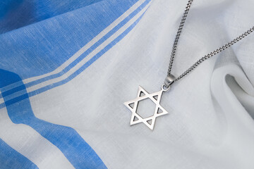 A silver-toned Star of David (Magen David) pendant on a chain rests on a softly draped blue and white striped fabric, clearly highlighting the Jewish religious and cultural symbol.