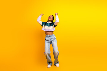 Happy young woman in colorful casual outfit celebrating with raised arms on a vibrant yellow backdrop full of energy