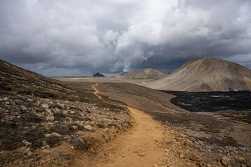 Trail across barren hills to Iceland Litli Hrútur volcano. Smoke from the active volcano rises on the horizon above a dark lava field under dramatic storm clouds
