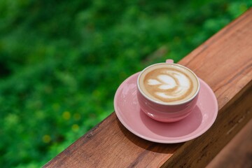 A pretty pink coffee cup with saucer holds a cappuccino or latte featuring perfect foam art, set on a warm wooden railing against a vibrant green, blurred natural background.