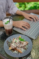 A person working remotely on a laptop while enjoying a sweet dessert (banana fritters/pisang cokelat) topped with cheese and a layered iced coffee drink at an outdoor cafe or balcony.
