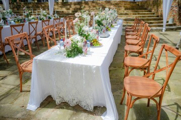 A beautifully set long table with a white lace tablecloth and fresh floral arrangements, surrounded by rustic wooden cross-back chairs at an outdoor wedding or garden party venue with stone stairs.