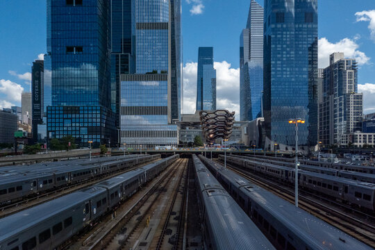 View of trains stretching into the distance beneath towering blue skyscrapers under a bright sky in Hudson Yard, New York City, New York, United States.