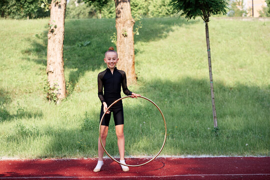A young gymnast performing with a hula hoop outdoors on a sunny day, showcasing athleticism and dedication. The lush greenery adds a vibrant and lively atmosphere to the scene. - Powered by Adobe