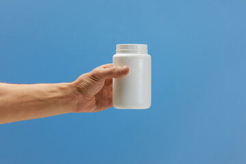 Hand holding white plastic bottle offering vitamins, supplements, or medication against a blue background