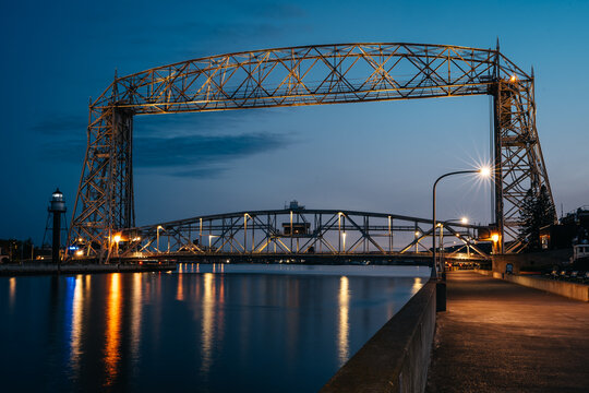 Aerial Lift Bridge illuminated at blue hour over the Duluth Ship Canal in Duluth, Minnesota, with reflections on calm water and the harbor lighthouse in the background.