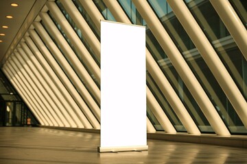 A blank white roll up banner stands in a modern building with angled window support structures behind it
